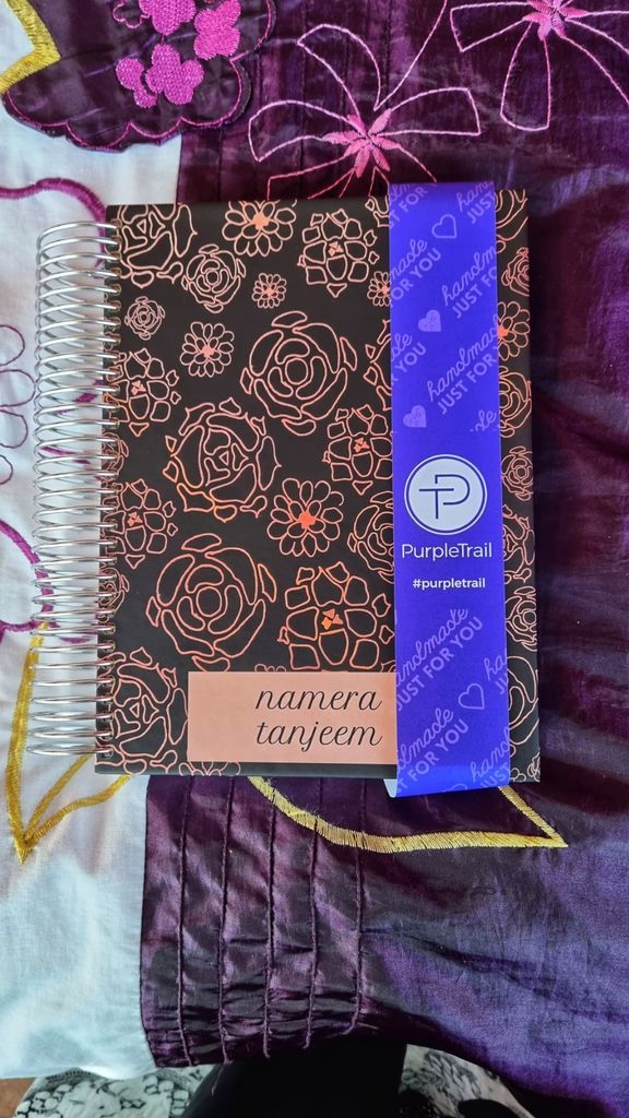 Photo of PurpleTrail Planner against a purple flowered fabric background. The cover has bronze flower patterns.