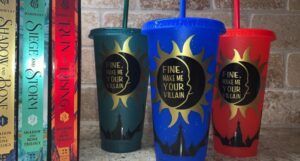 image of three plastic tumblers bearing the words "Fine, Make Me Your Villain" in gold text. Tumblers are red, blue, and green. To the left are the books in Leigh Bardugo's Shadow and Bone series
