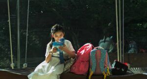 photo credit: Tam Wai. ID: a young Asian girl wearing a white dress with a blue belt sits in reading a book; there are two backpacks next to her on the bench she is sitting on, and the background is dark with a suggestion of trees and a brick wall.