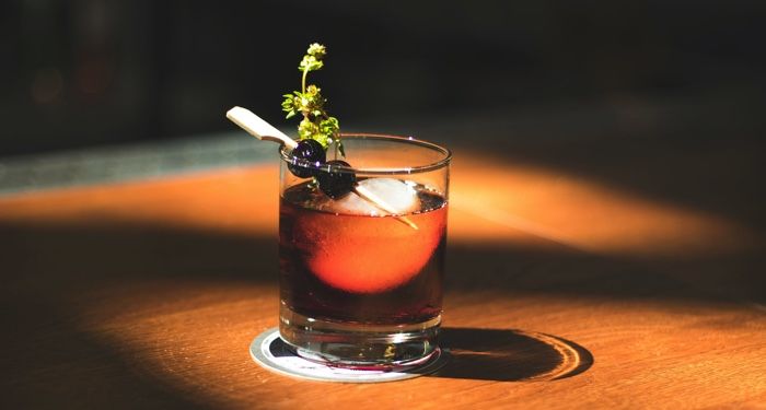 an old fashioned cocktail with a cherry, herb garnish, and spherical ice
