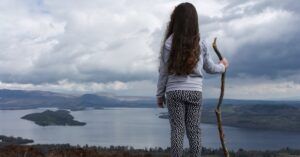 little girl standing on a hilltop overlooking the ocean with a walking stick for survivalist outdoors nature feature