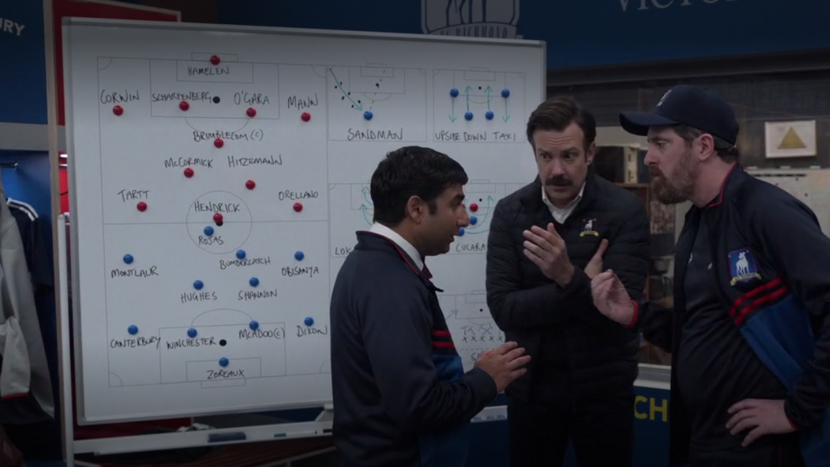 Nate, Ted, and Beard huddle in front of a whiteboard with their plays drawn on it