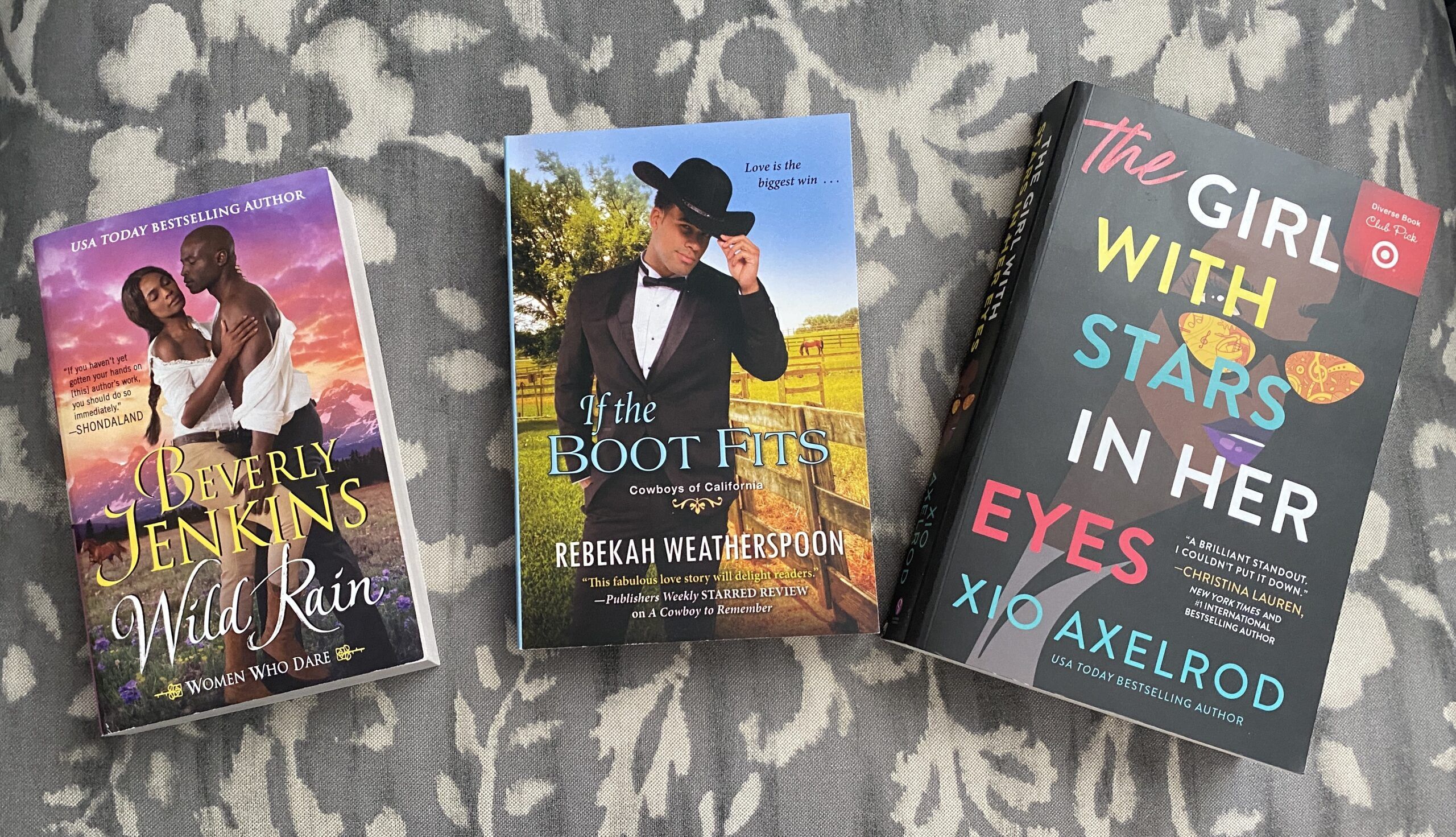 Photograph of three paperbacks on a paisley background. The book on the left is a mass market paperback, Wild Rain by Beverly Jenkins. The book in the middle is a mass max, If the Boot Fits by Rebekah Weatherspoon, and the book on the right is a trade paperback, The Girl With Stars in Her Eyes by Xio Axelrod. The three increase in size from left to right. 