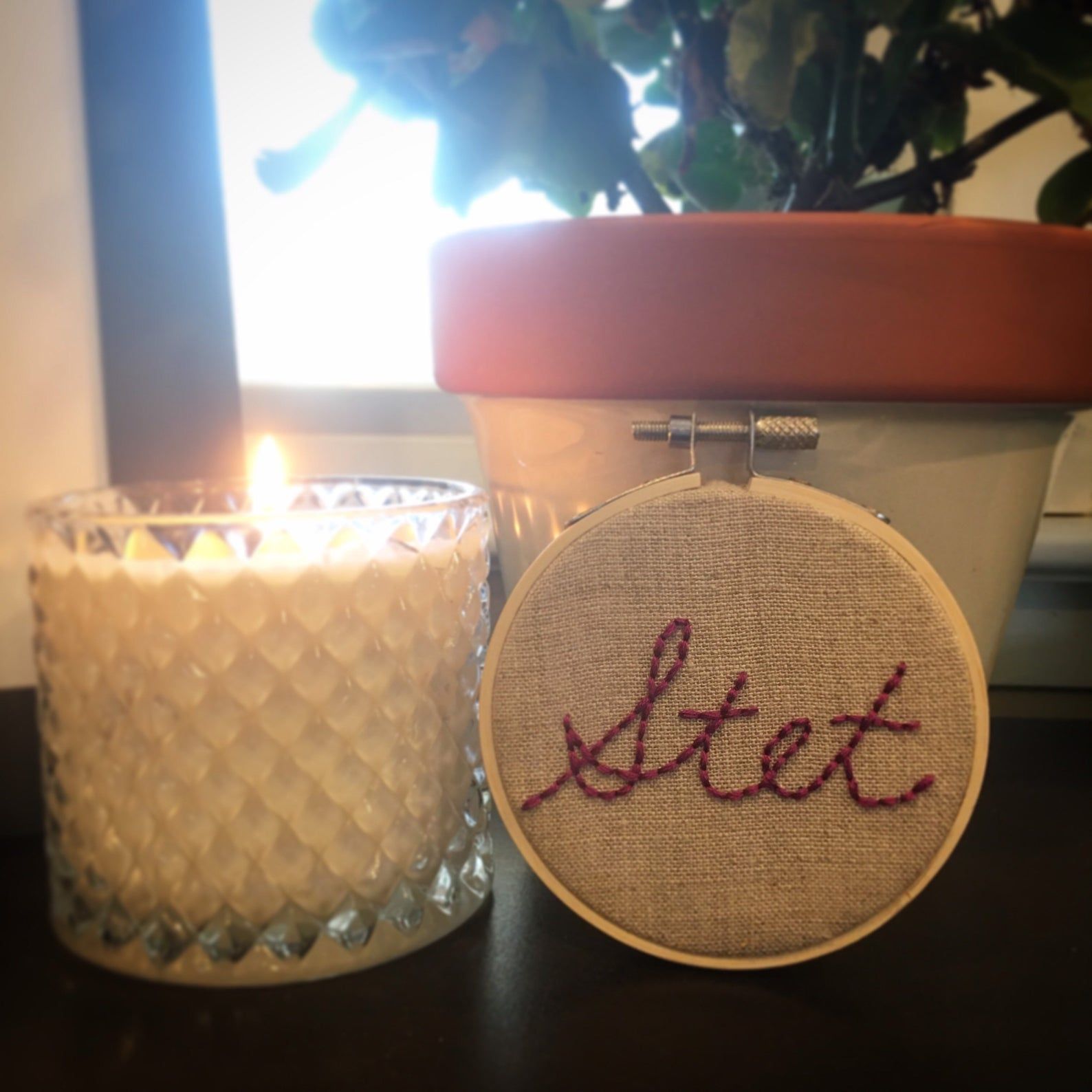 A small embroidery of the abbreviation "Stet" in cursive. The embroidery hoop is resting against a plant pot and is beside a burning candle.