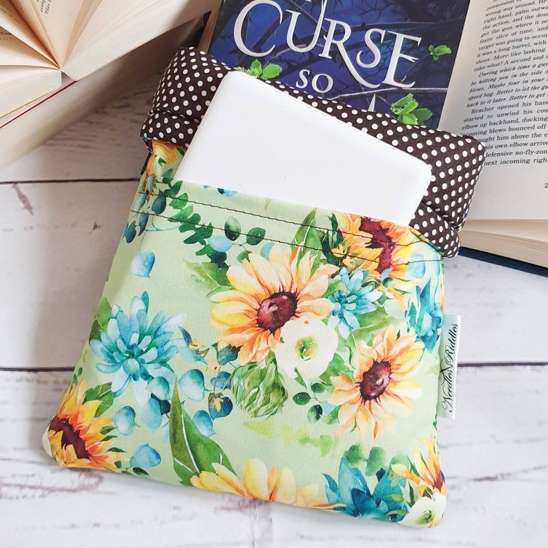 A book sleeve with a book and kindle peeking out. The book sleeve is light green with yellow, blue, and white flower patterns. 