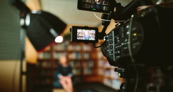 person sitting in front of a camera on a TV or film set; shelves of books are in the background https://unsplash.com/photos/KieCLNzKoBo