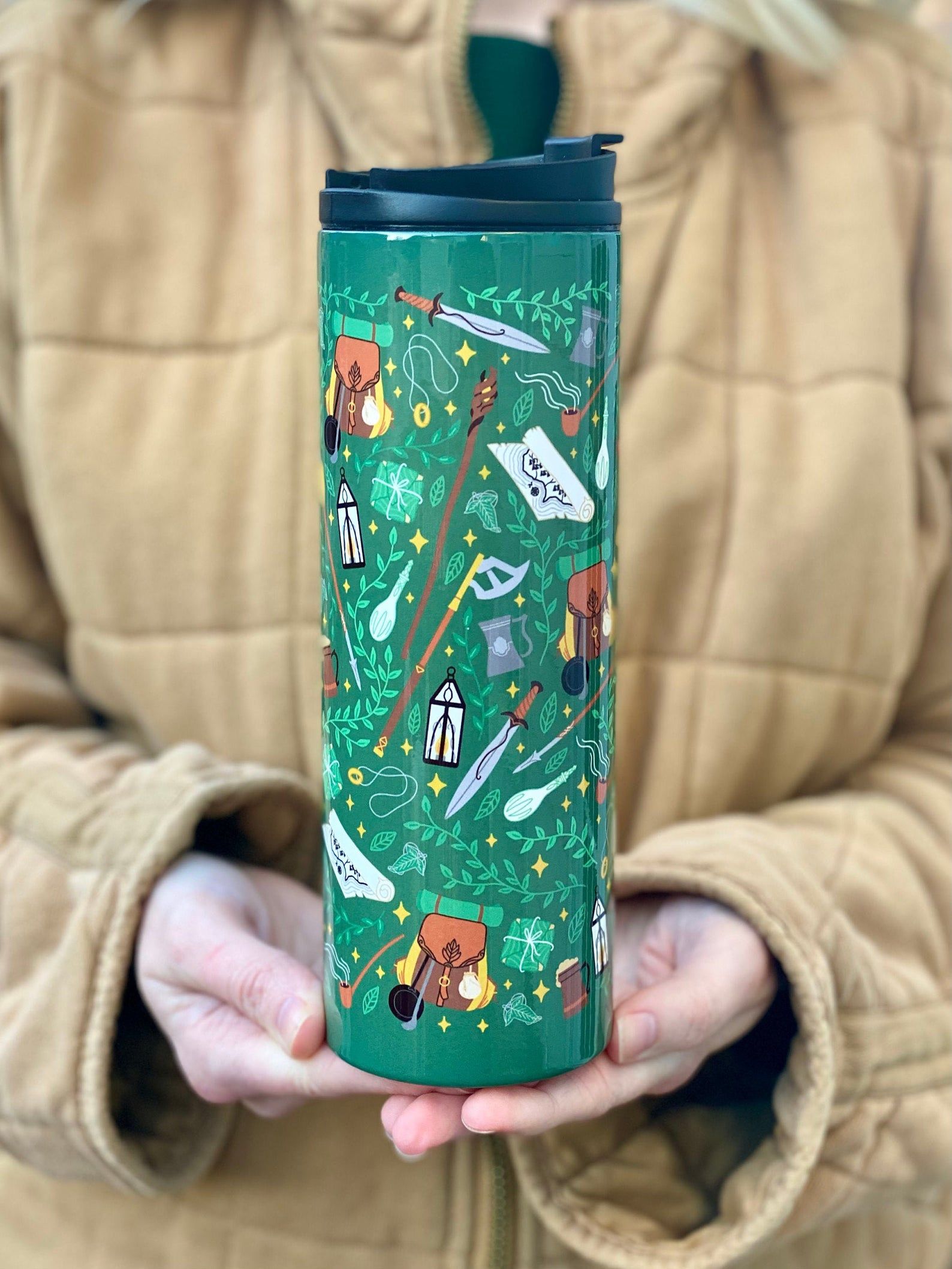 A person holding a tumble in their hands. The tumbler is green with a pattern of leaves, swords, backbacks, and weapons. 