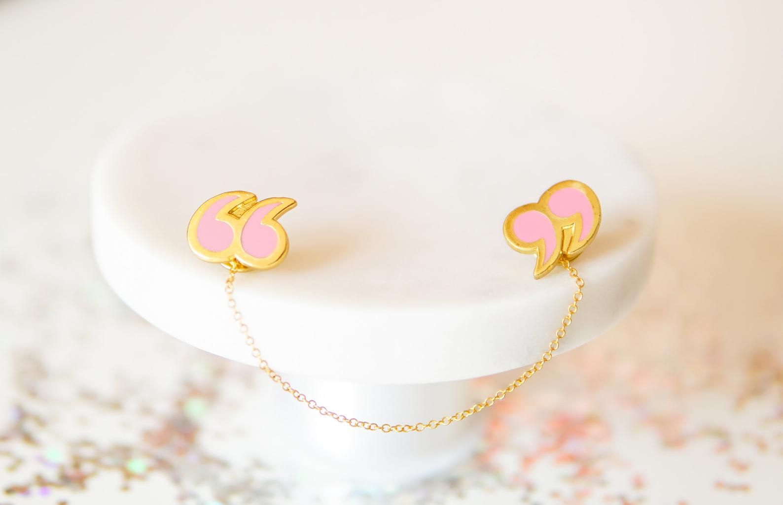 Two pink and gold quotation mark pins, linked by a gold chain. They are resting on a white stand.