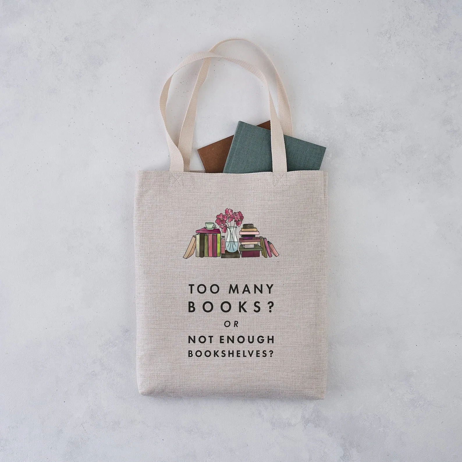 A tote bag with books peeking out rests on a gray background. The tote is light brown in colour and has an image of many books side-by-side and stacked up with a flower vase in the middle. Below, it reads: Too many books? Or not enough bookshelves? 