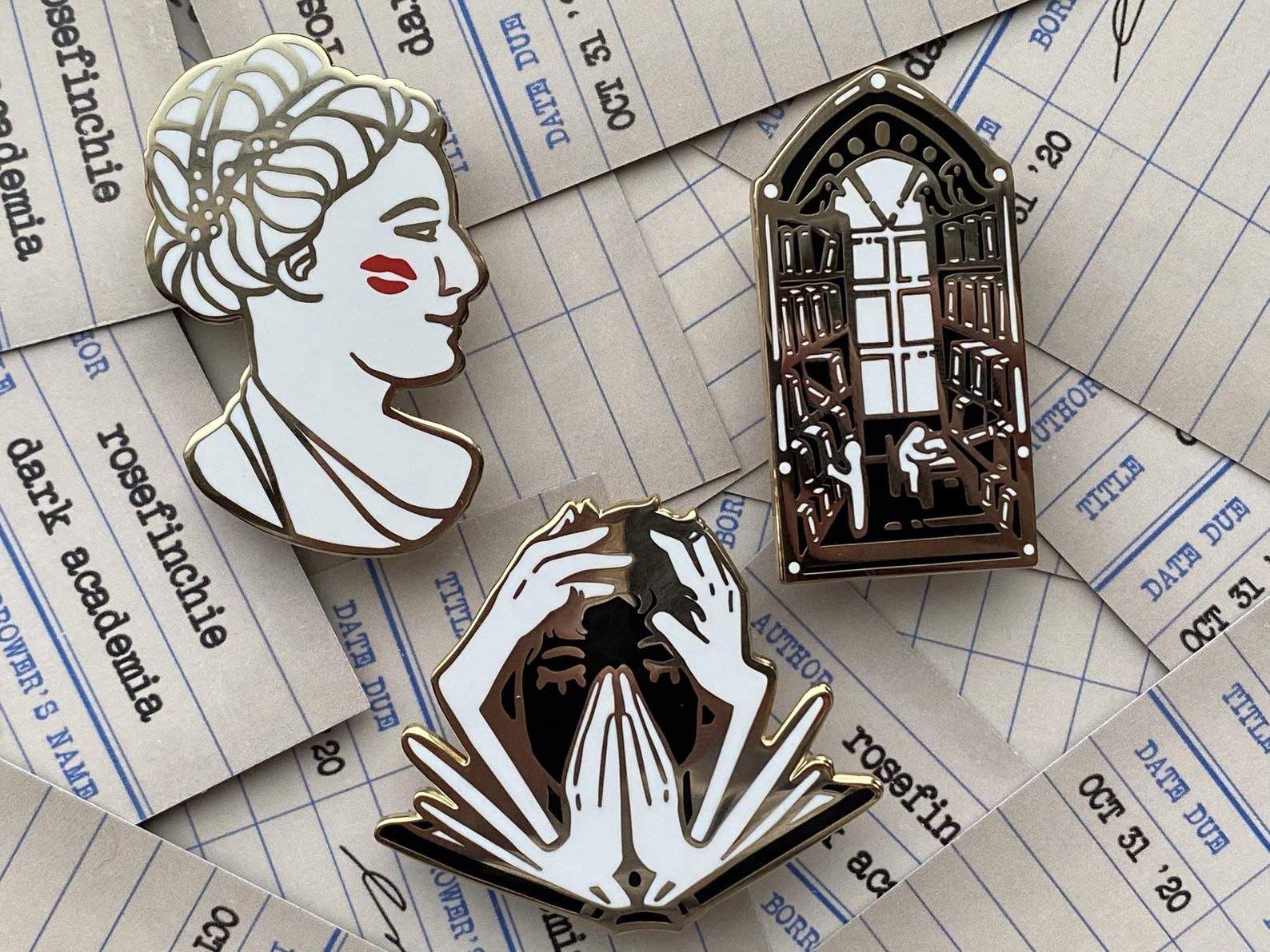 three dark academia enamel pins against a backdrop of library due date cards