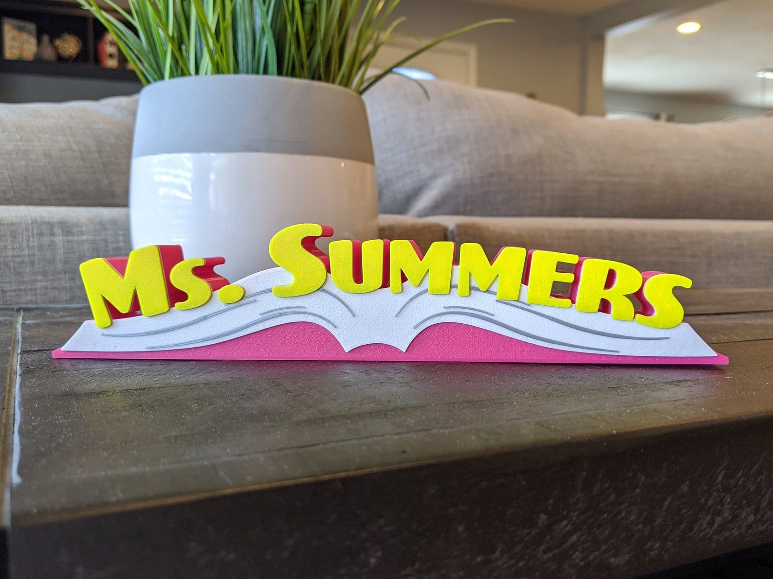 An image iof a name plate sitting on a wooden table with a couch and plant in the background. The name plate is in the shape of a pink open book, and the name "Ms. Summers" appears in yellow text on the book.