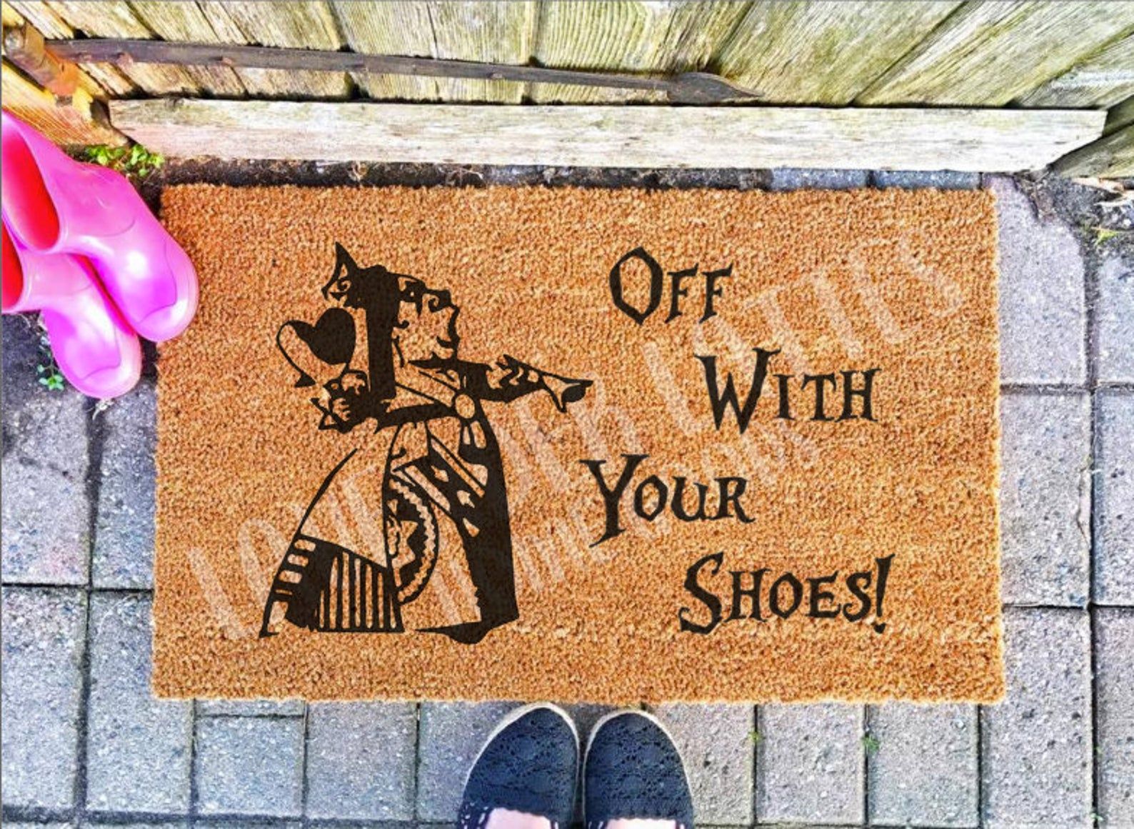 Image of a brown doormat with an image of Alice in Wonderland's Queen of Hearts. Text reads "Off With Your Shoes!"