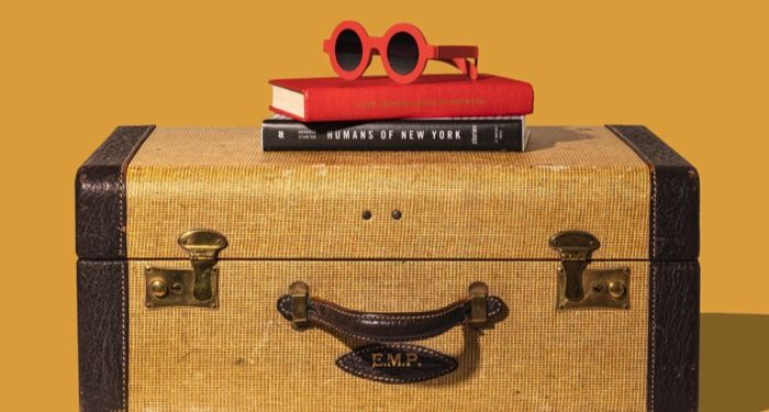 a brown and mustard suitcase with a stack of books and bright red plastic sunglasses on top