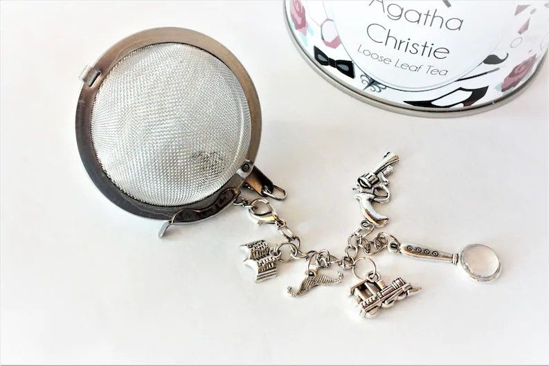 ball style tea infuser with charms of an open book, handlebar mustache, train, magnifying glass, and gun, all silver.