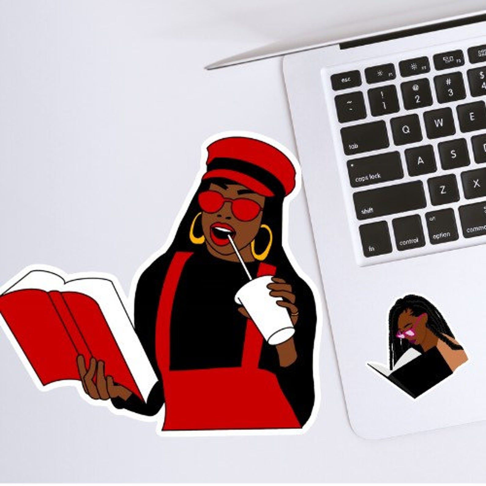 Picture of a vinyl sticker next to a silver laptop. The sticker is of a stylish Black woman with long hair. She is wearing a red hat, a black shirt and red jumper, red sunglasses, red lipstick, and gold hoops. She's reading a red book and drinking from a white cup with a straw.