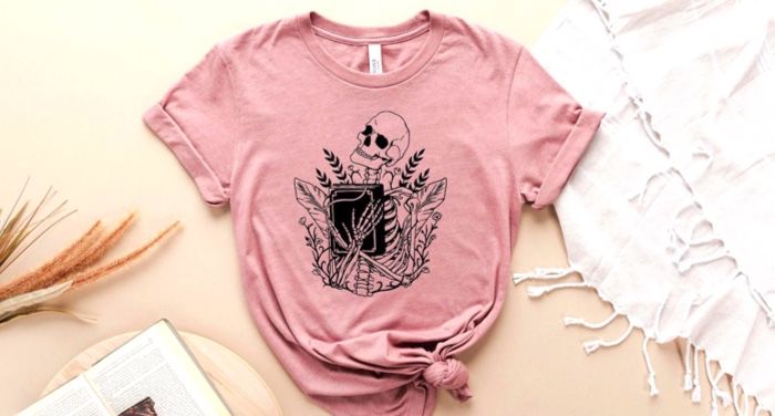 Pink t-shirt depicting a black line art drawing of a skeleton clutching a black book to its chest with a background of plants.