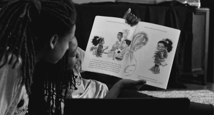 grayscale photo of two people reading a picture book in front of a laptop computer