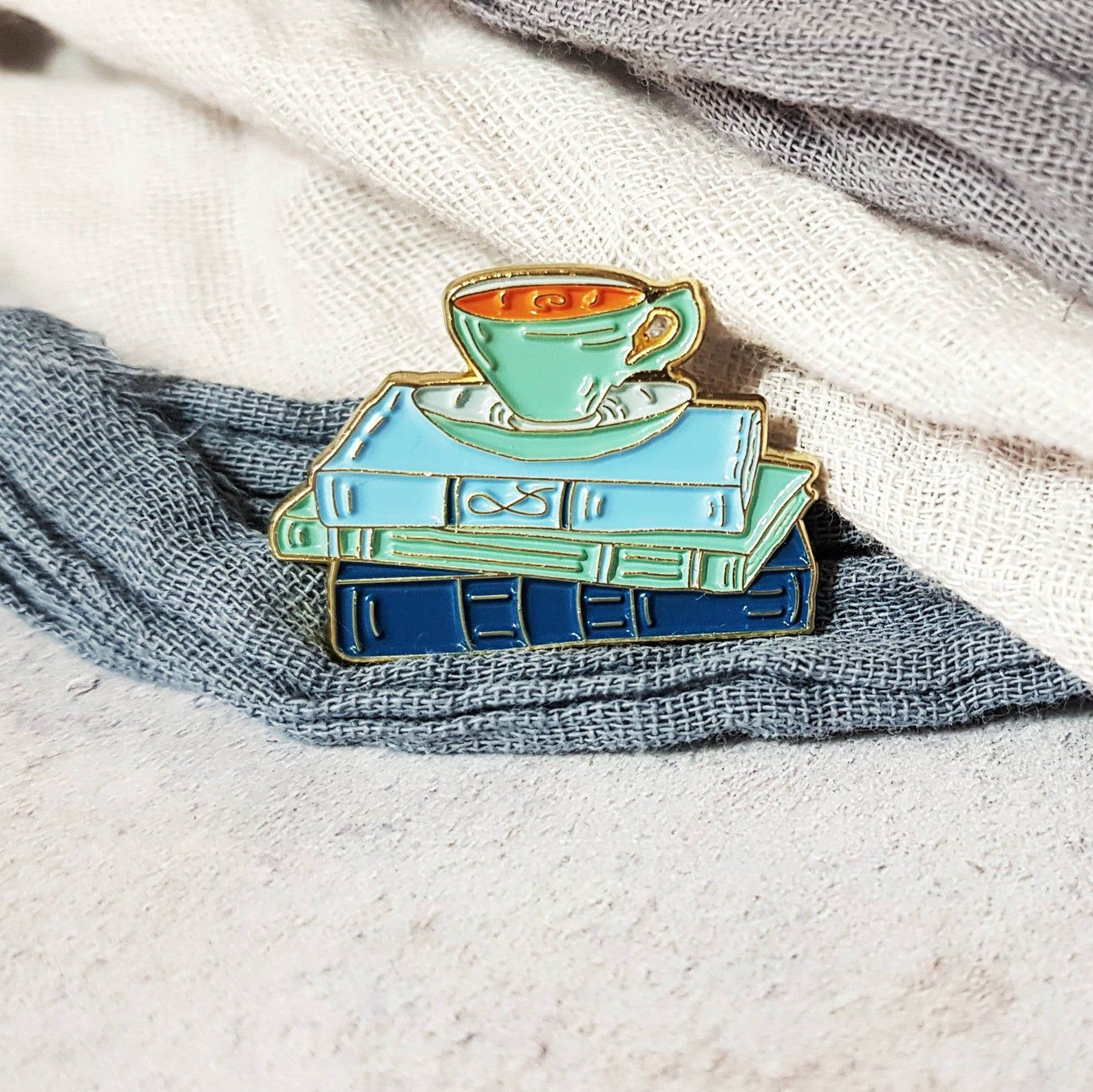 a blue and green enamel pin depicting a stack of books and a tea cup and saucer.