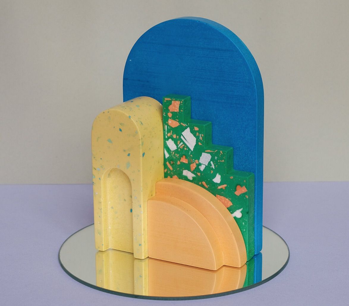 Sculpture-style bookend, with a small orange terrazzo arch, a mid-size yellow terrazzo arch, a green terrazzo stair step, and a big blue background. 