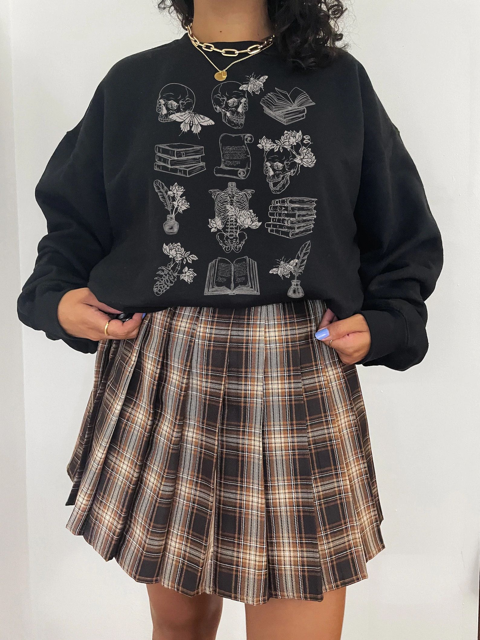 Photo of woman wearing a black sweatshirt with line drawings of skulls, books, flowers, and other imagery associated with the subgenre of dark academia.