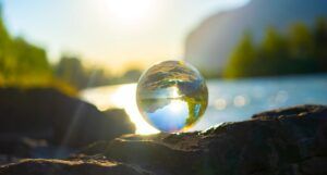 a photo of a clear marble positioned in front of a beautiful lake with mountains and trees in the background; the marble is reflecting and flipping the surroundings and it looks like a tiny planet earth