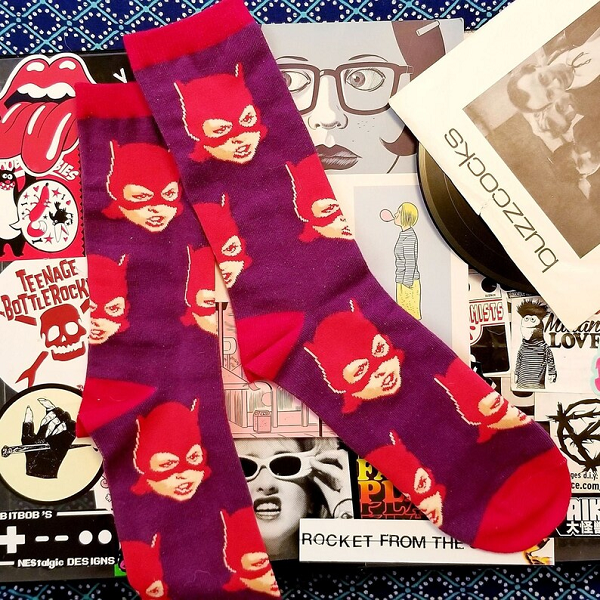 A pair of red and purple socks with a girl in a Batman mask printed on them, laid over a collage of Ghost World memorabilia.