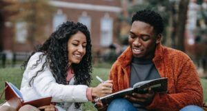 two young college-aged adults sitting in front of a brick building comparing notes