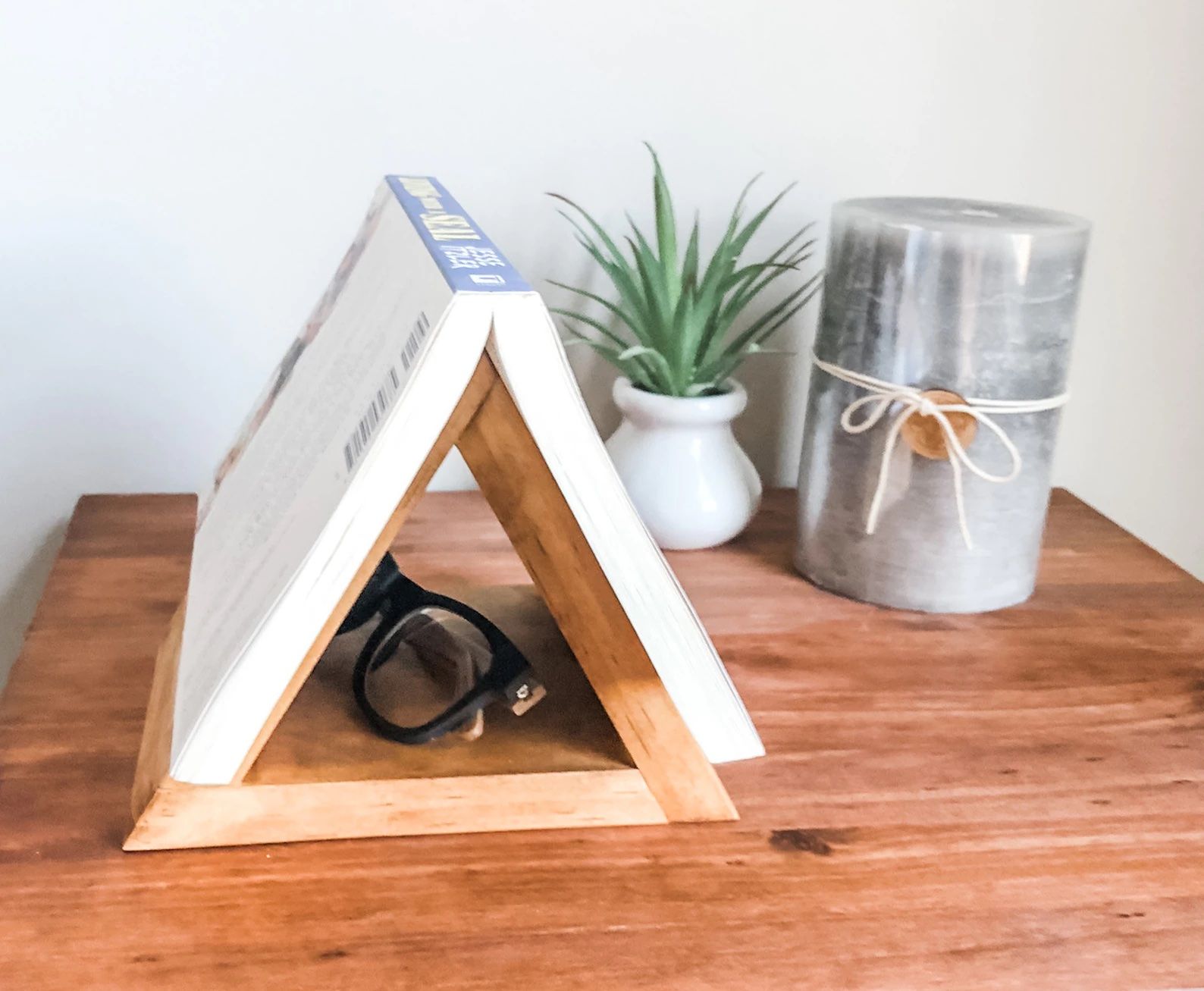 A triangular book holder sits on a nightstand, and a book is set down, open, on top of the holder so the pint of the triangle holds the book open. It's displayed on a nightstand with a candle and plant.