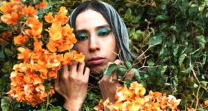 a trans woman surrounded by green plants holding yellow flowers