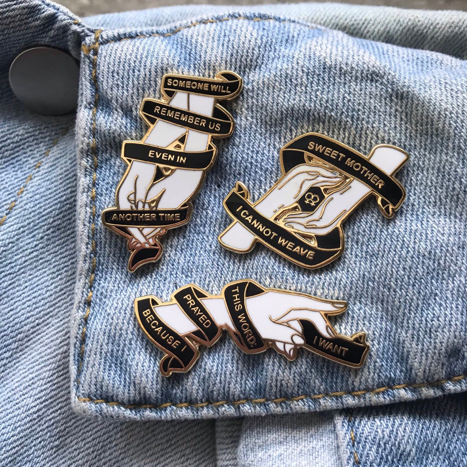 A set of three enamel pins on a denim jacket. The pins depict hands holding or reaching, with black ribbon running around them and lines from Sappho in gold.