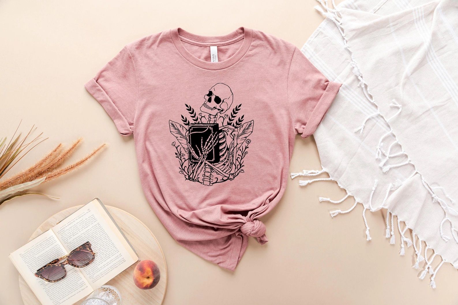 Pink t-shirt depicting a black line art drawing of a skeleton clutching a black book to its chest with a background of plants.