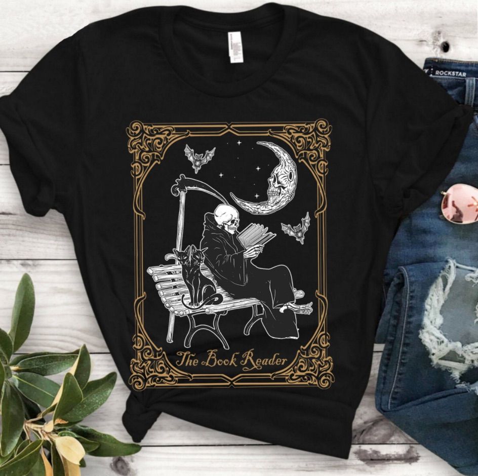 Image of a black tee shirt. In the center is a skeleton seated on a bench reading a book. He's wearing a black cloak, sitting beside a double-headed black cat. Behind him is a scythe. 