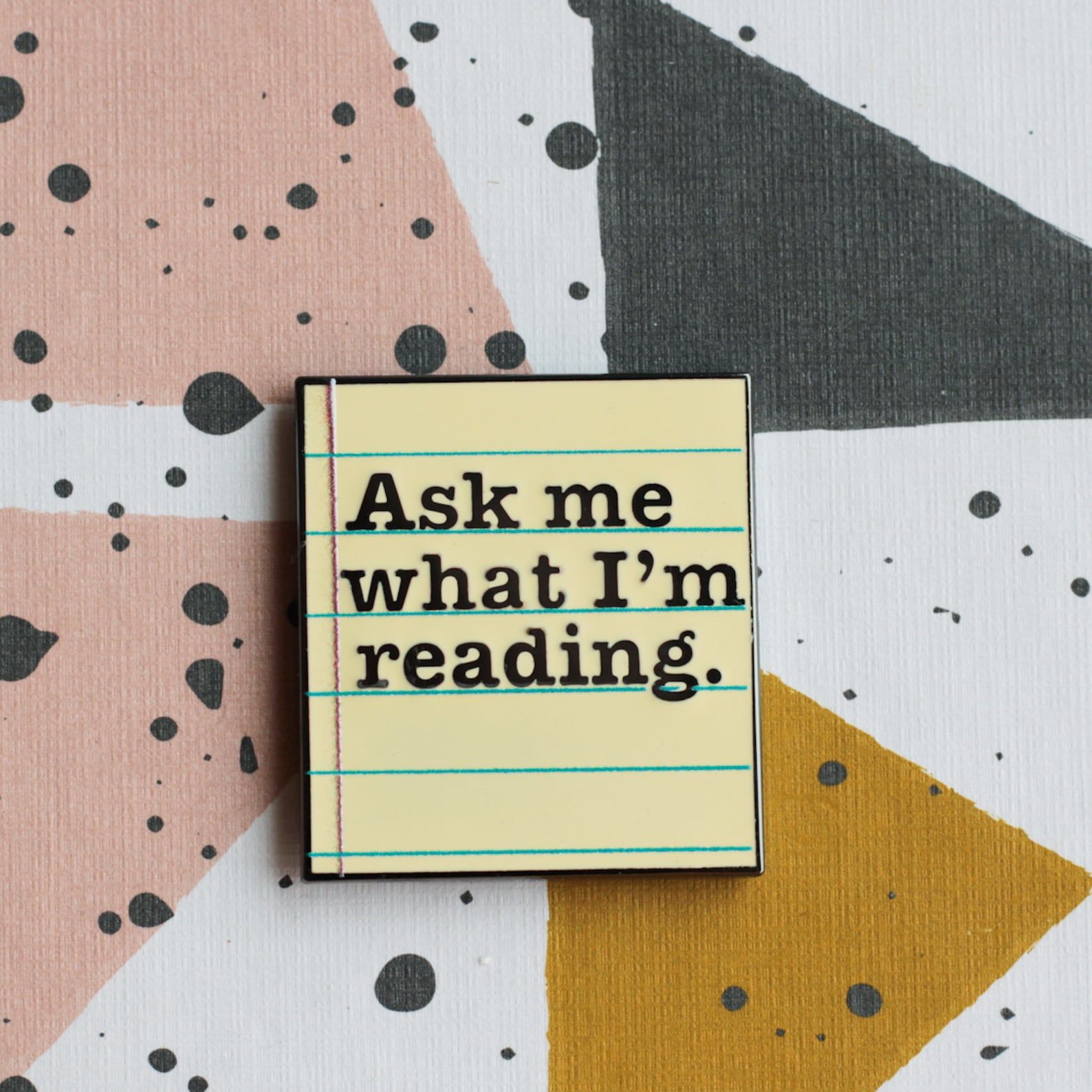 An enamel pin that looks like a piece of lined paper and reads "Ask me what I'm reading."