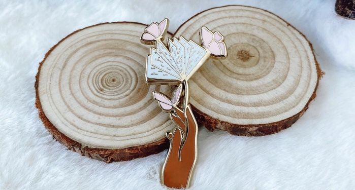 An enamel pin depicting a brown hand holding an open book stylized as a flower, with pink butterflies