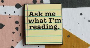 An enamel pin that looks like a piece of lined paper and reads "Ask me what I'm reading."