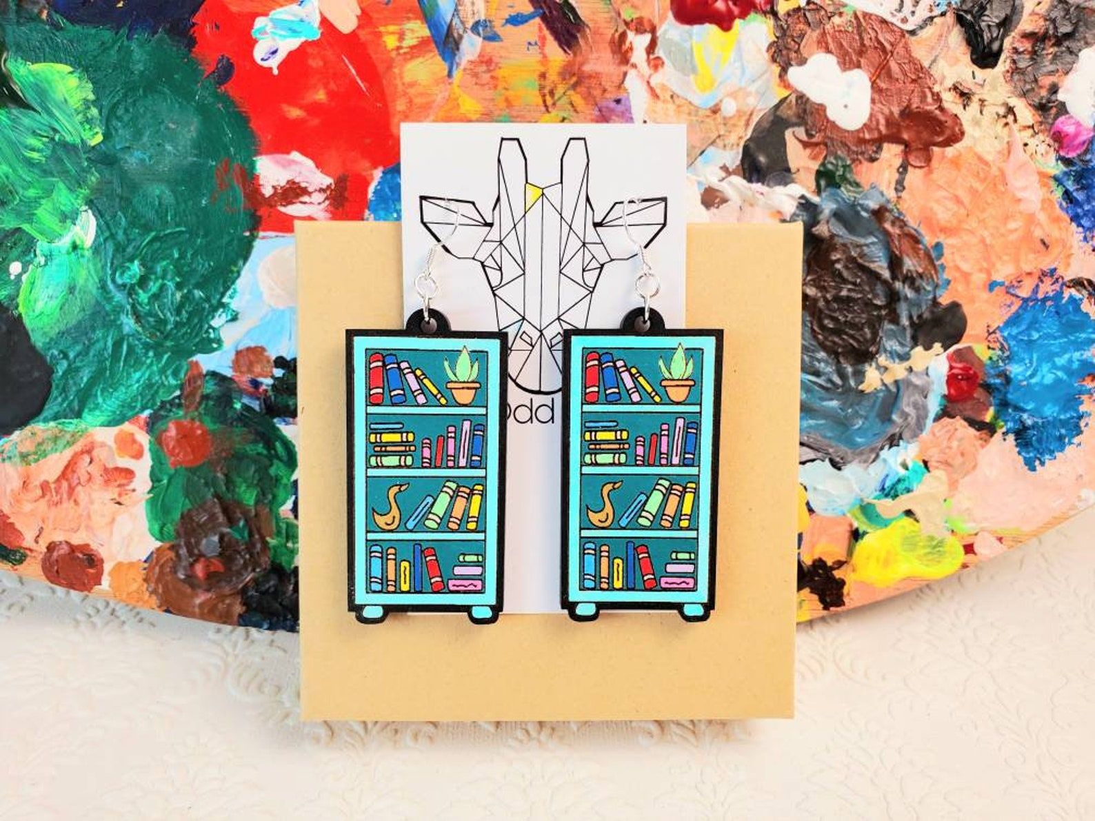 Dangly teal earrings in the shape of bookshelves filled with colorful books and plants.