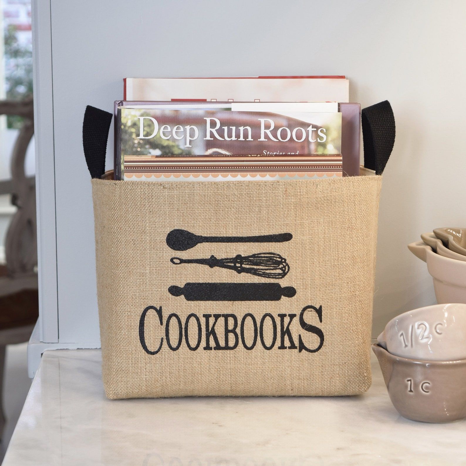 Image of a canvas bin. The text reads "cookbooks" and above it are a rolling pin, whisk, and spoon. 