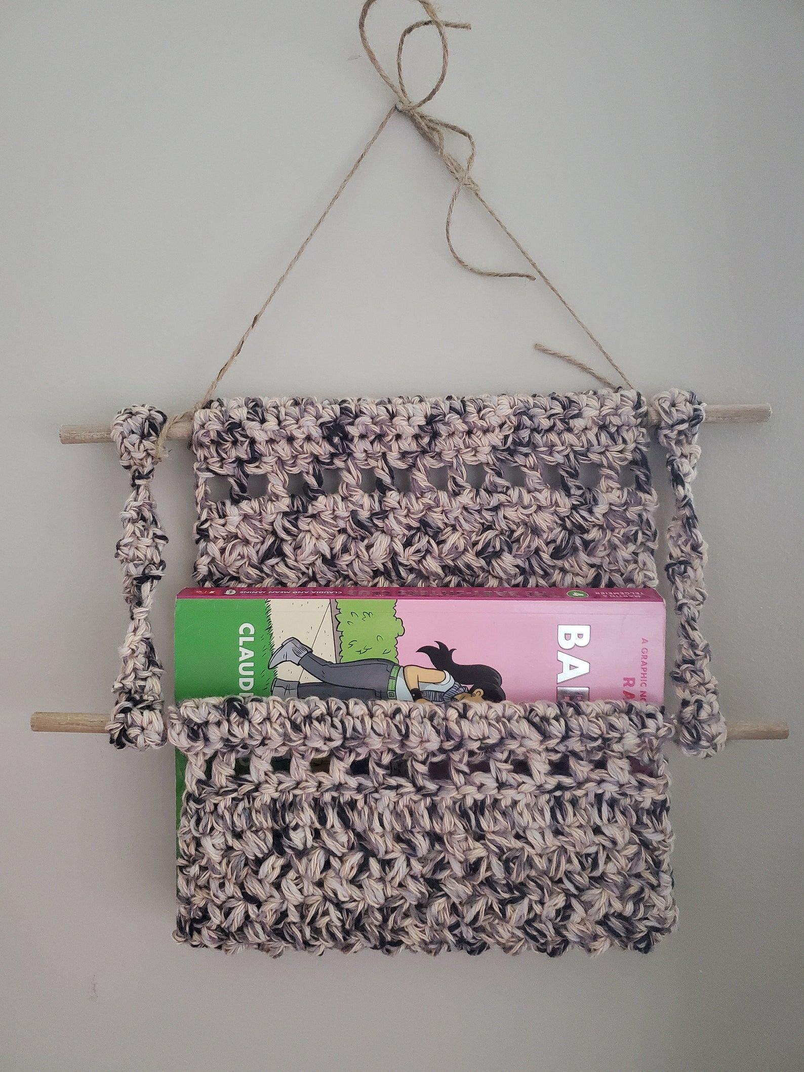 Purple pink and black yarn are crocheted into a wall hanging with a slot for holding a book. 