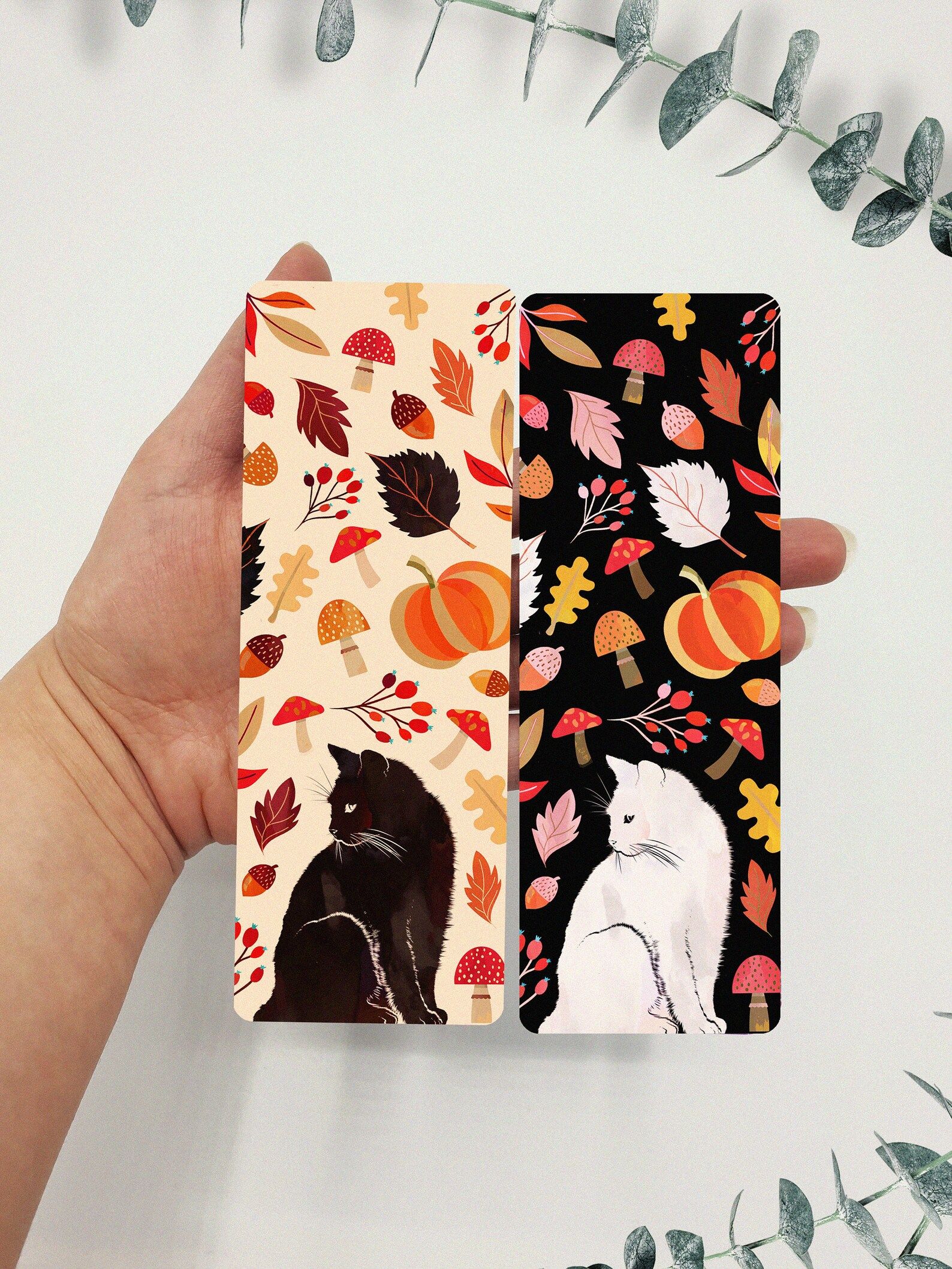 Image of two bookmarks being held by a white hand. The bookmark on the left features a black cat on a white background, with mushrooms and leaves surrounding it. The bookmark on the right is the same, except the cat is white and background is black. 