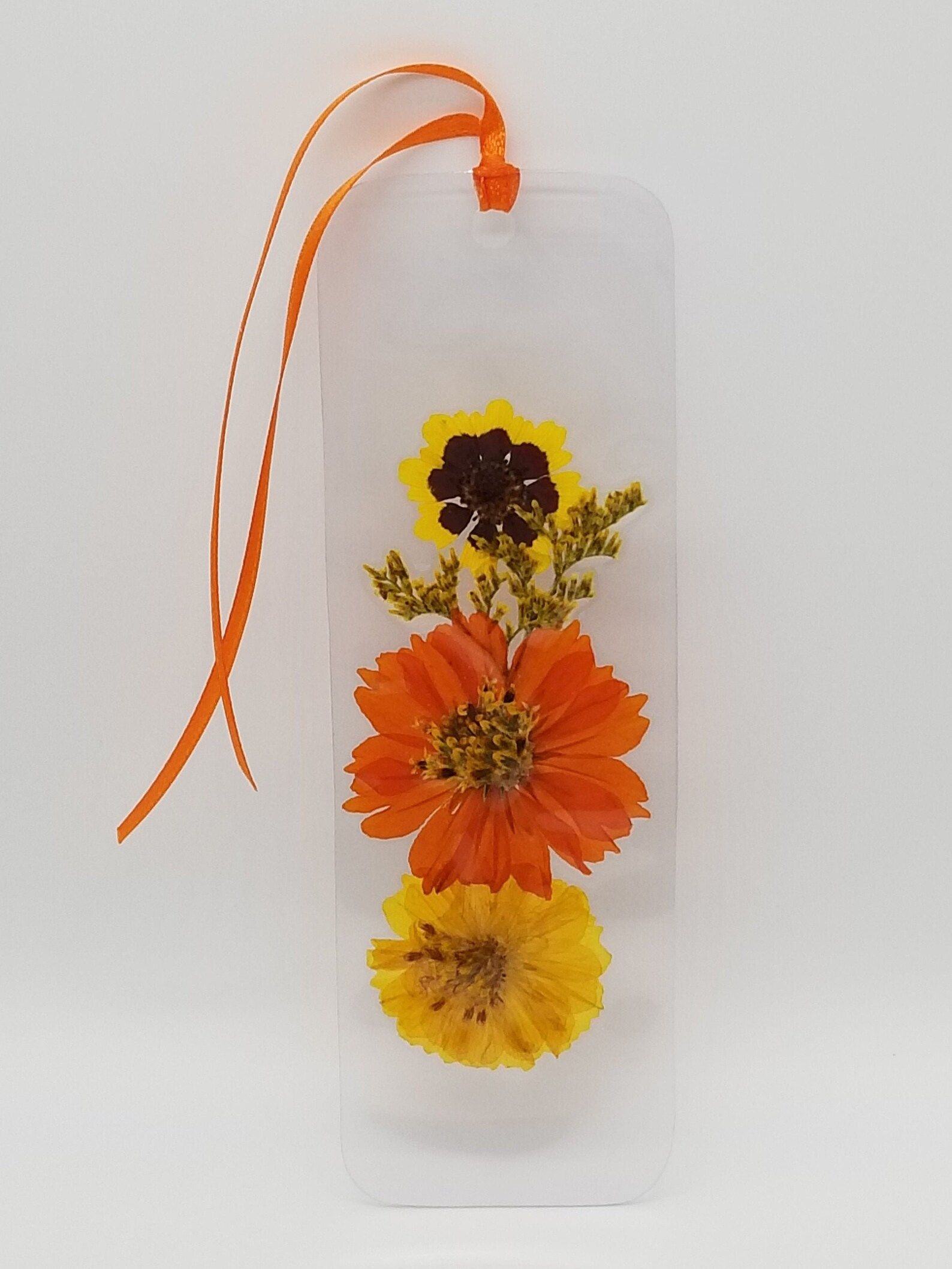 Image of pressed flower bookmark. Inside the bookmark are two yellow flowers and one orange flower. It has an orange tassel.