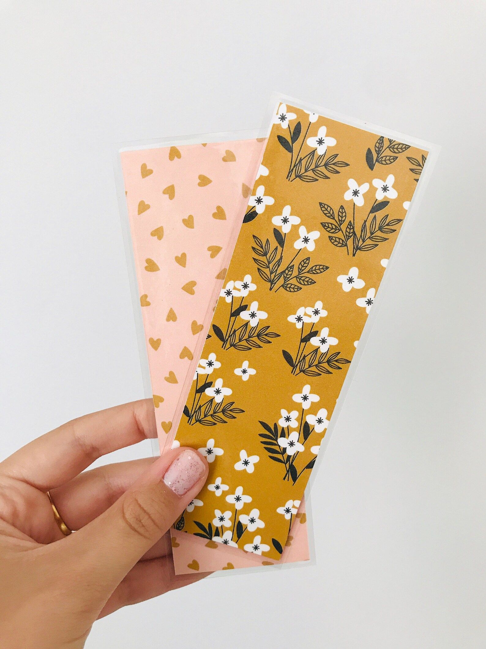 Image of a white hand holding a bookmark. The front of the bookmark at the foreground is golden yellow with white flowers. The back of the bookmark, in the background, is pink with gold hearts. 