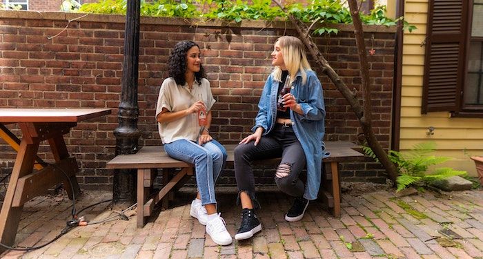 Image of two teen girls sitting on a bench. One is brown skinned and one is olive skinned.