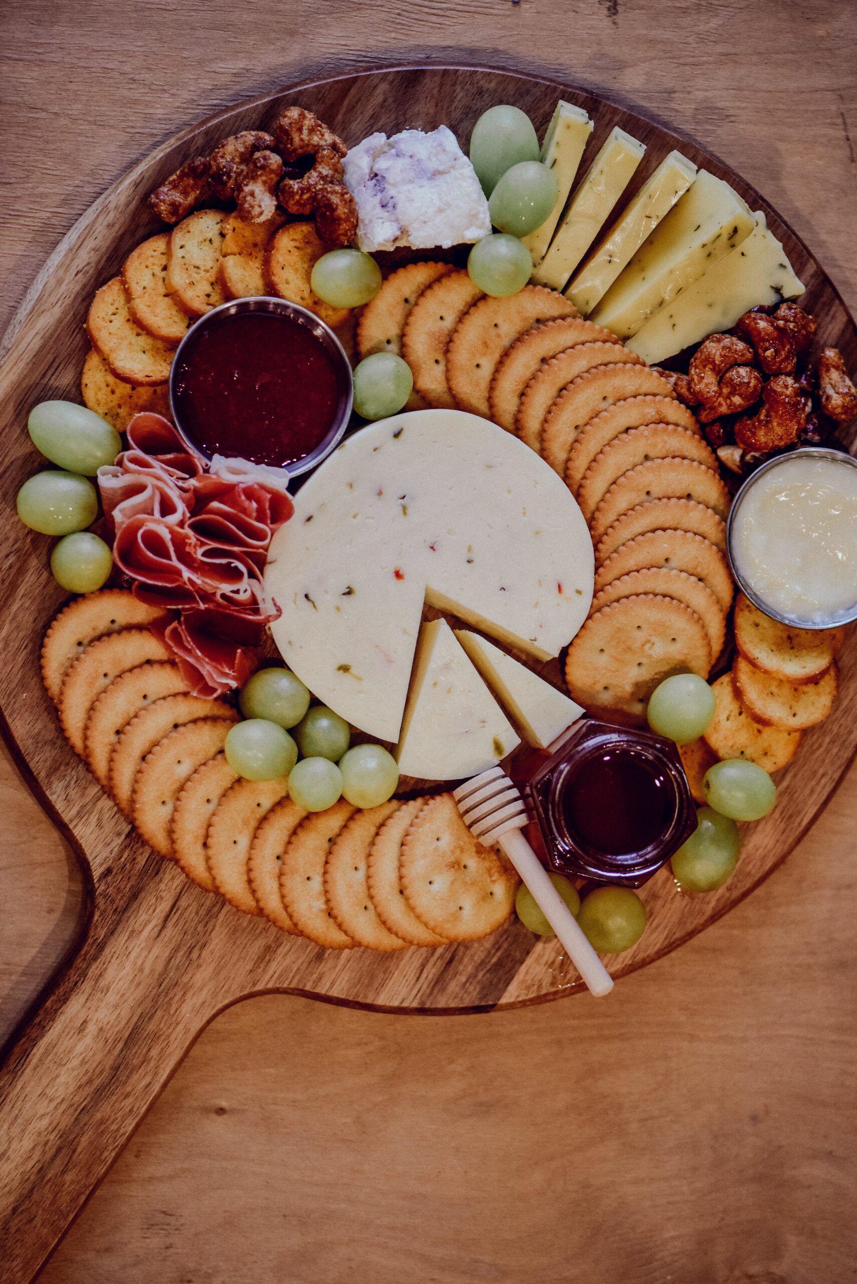 cutting board with handle holding crackers, jam, cheese, meat, and fruit