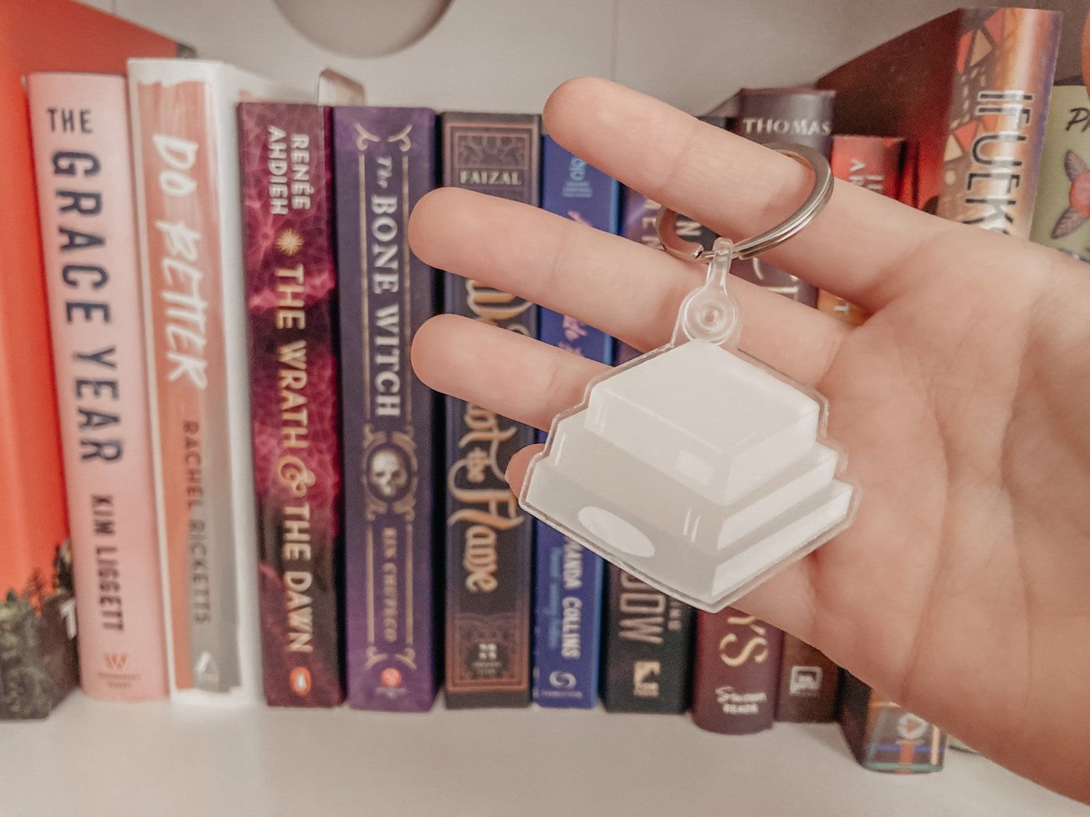 Ombre ivory book stack keychain held by a white hand in front of a bookshelf.