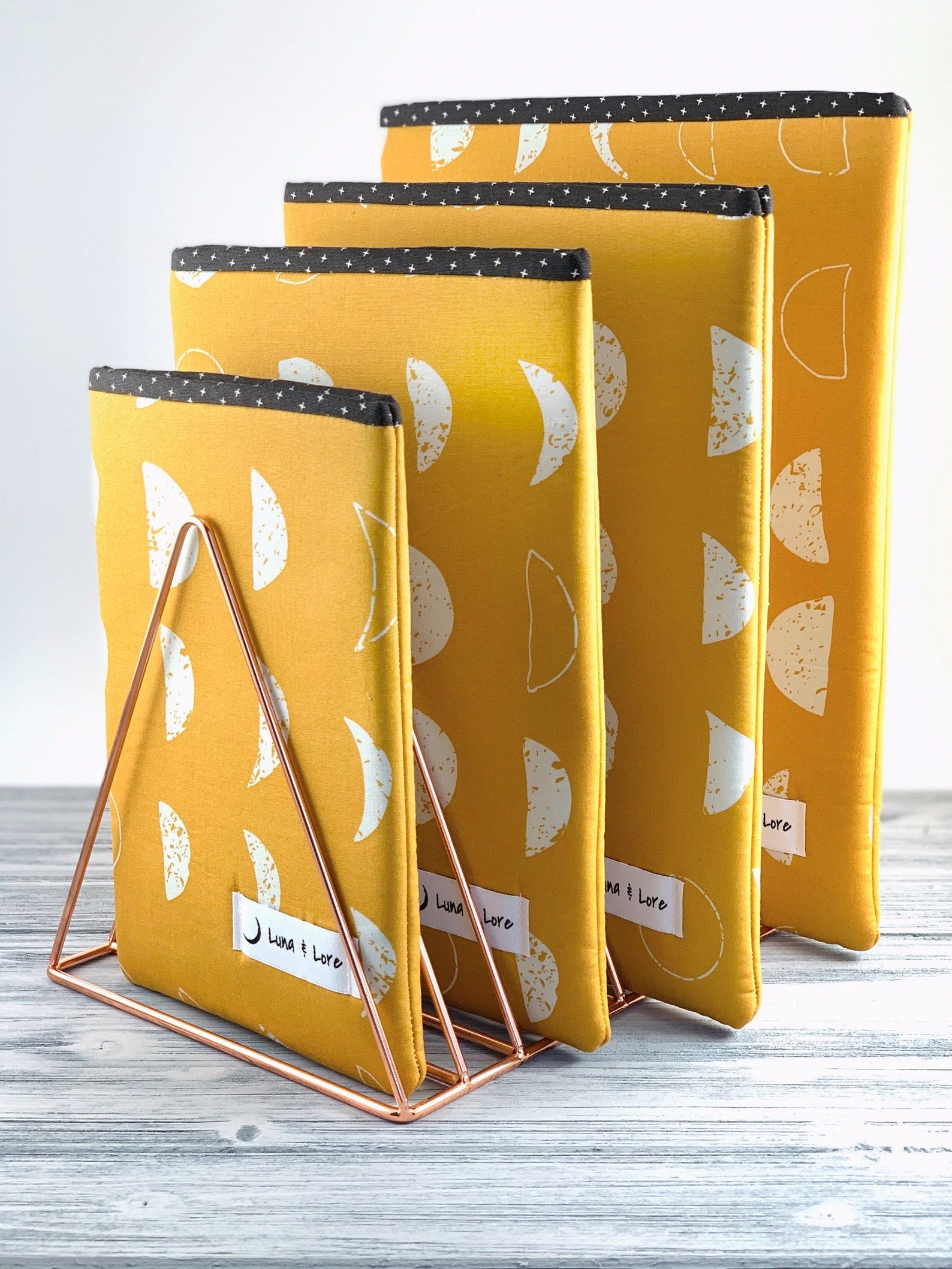 Four mustard-color book sleeves, each featuring the moon phases, standing up in a metal bookstand. 