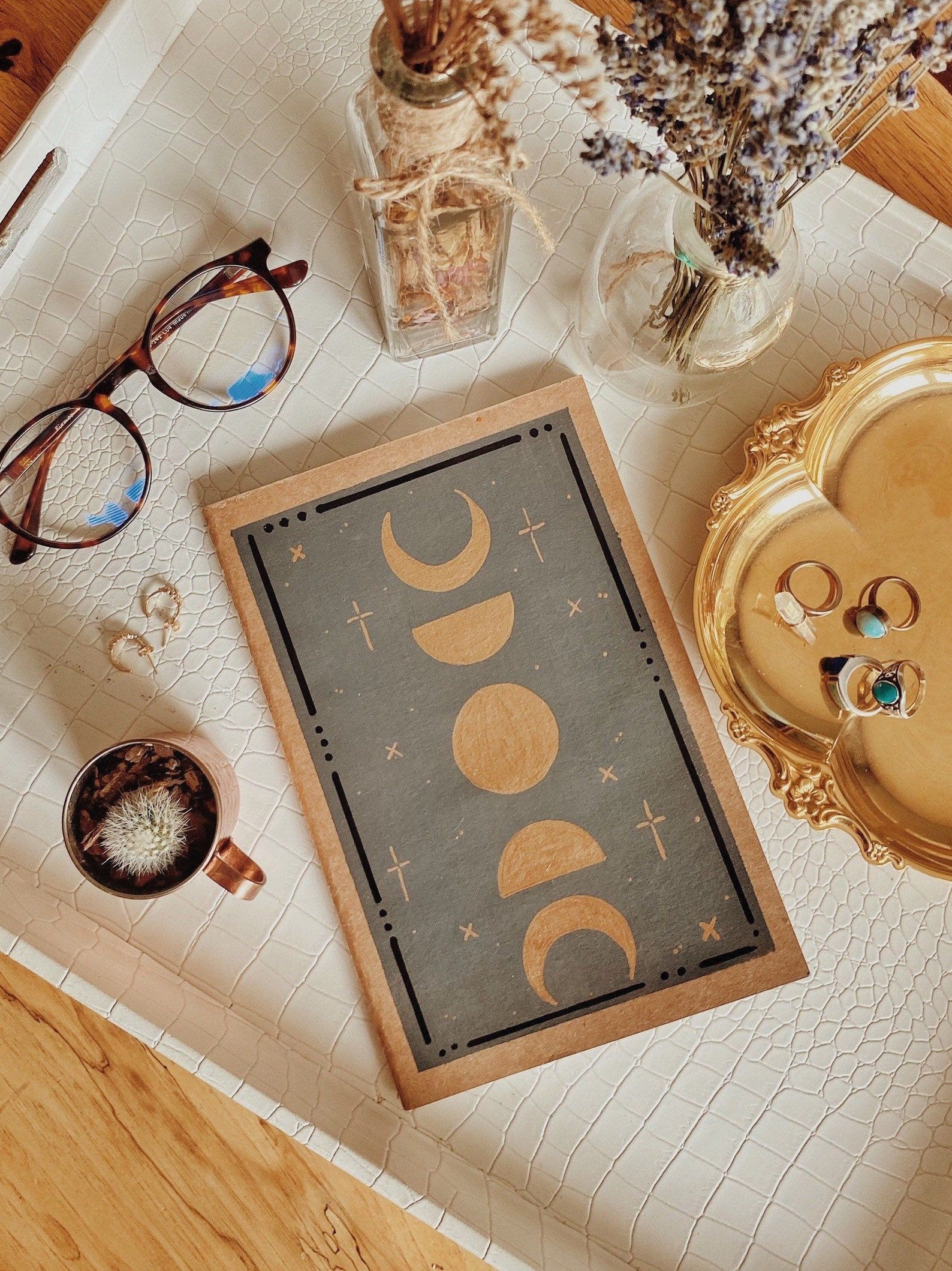 Image of a journal with moon phases on the cover. It's on a tray with jewelry, a cactus, and eye glasses. 