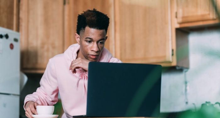 a Black person looking at their laptop computer while holding a cup of coffee in one hand