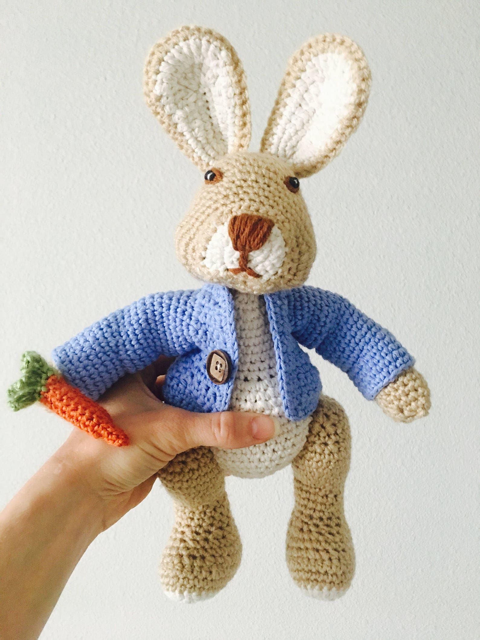A crocheted Peter Rabbit stuffed toy, wearing a blue cardigan and holding a carrot.