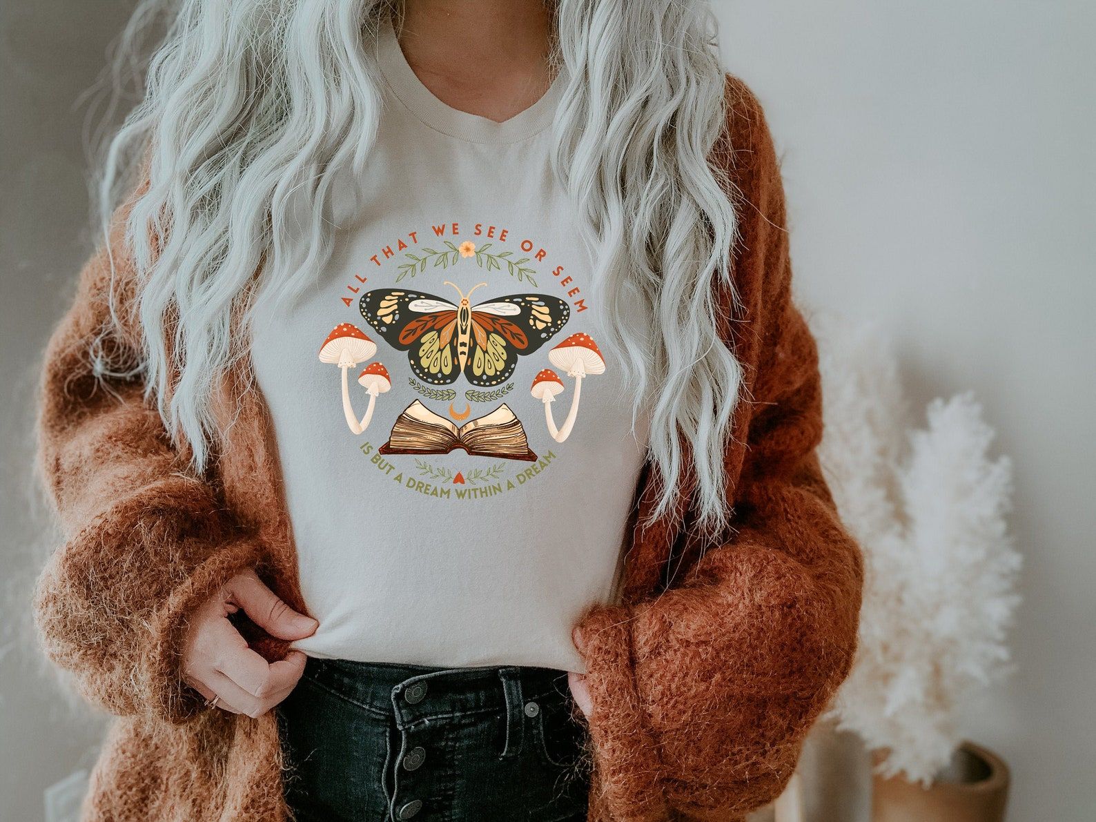 Image of a gray shirt with a butterfly, book, and toadstools. The quote on the shirt reads All that we see or seem is but a dream within a dream.