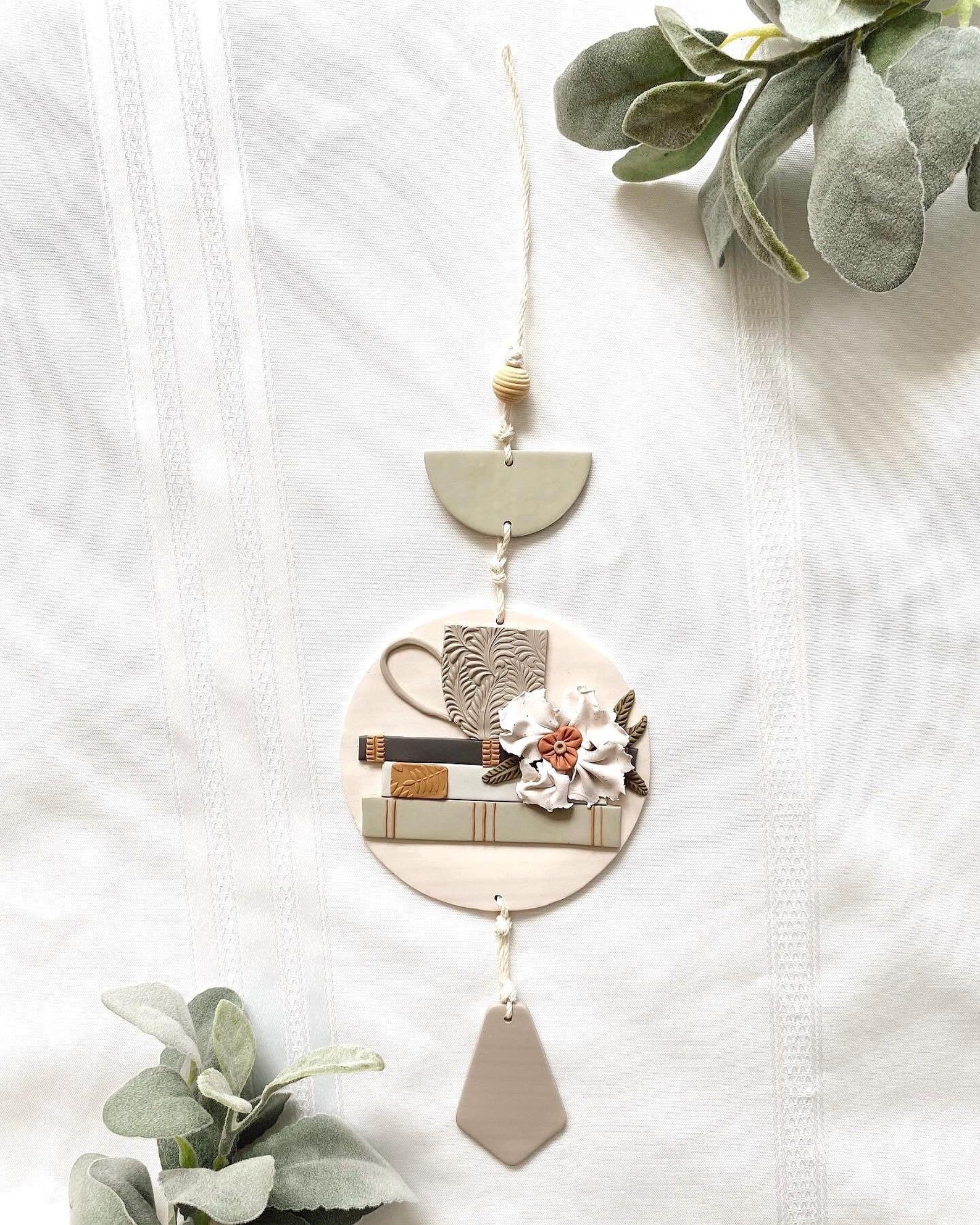 Polymer clay wall hanging, featuring a taupe mug on a stack of neutral books, with a white flower to the side. 